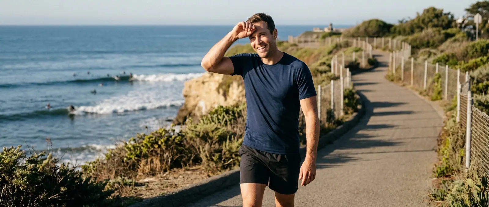 A man in a navy tee smiles on a coastal path above the surf